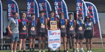 Group of boys in gray and black cross‑country uniforms wearing medals, posing with a banner that says CROSS COUNTRY CHAMPION behind them.