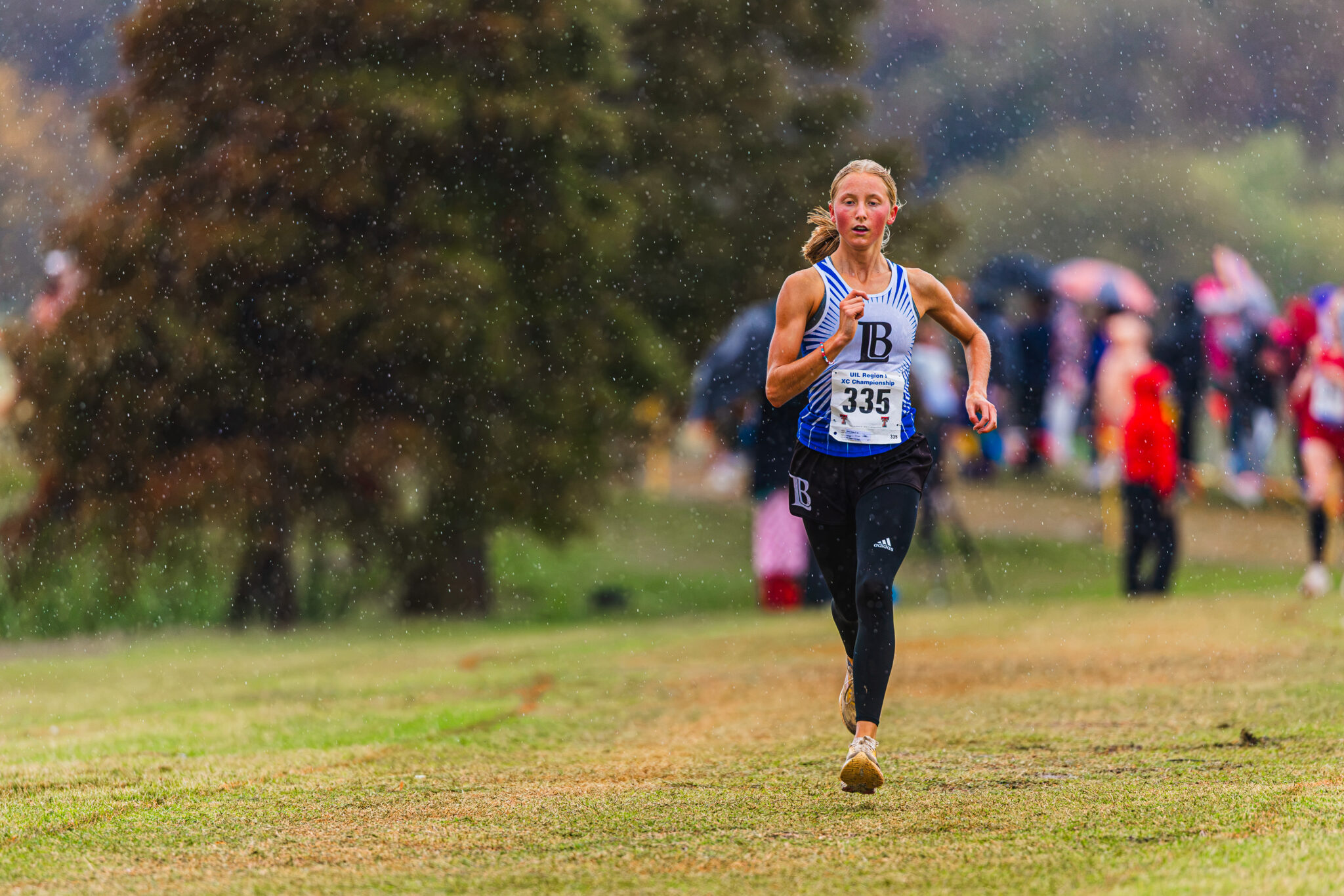 DE3_7087 | Press Pass Sports Female runner in a blue-white kit with bib 335 racing on a grassy field in the rain, spectators in the blurred background.