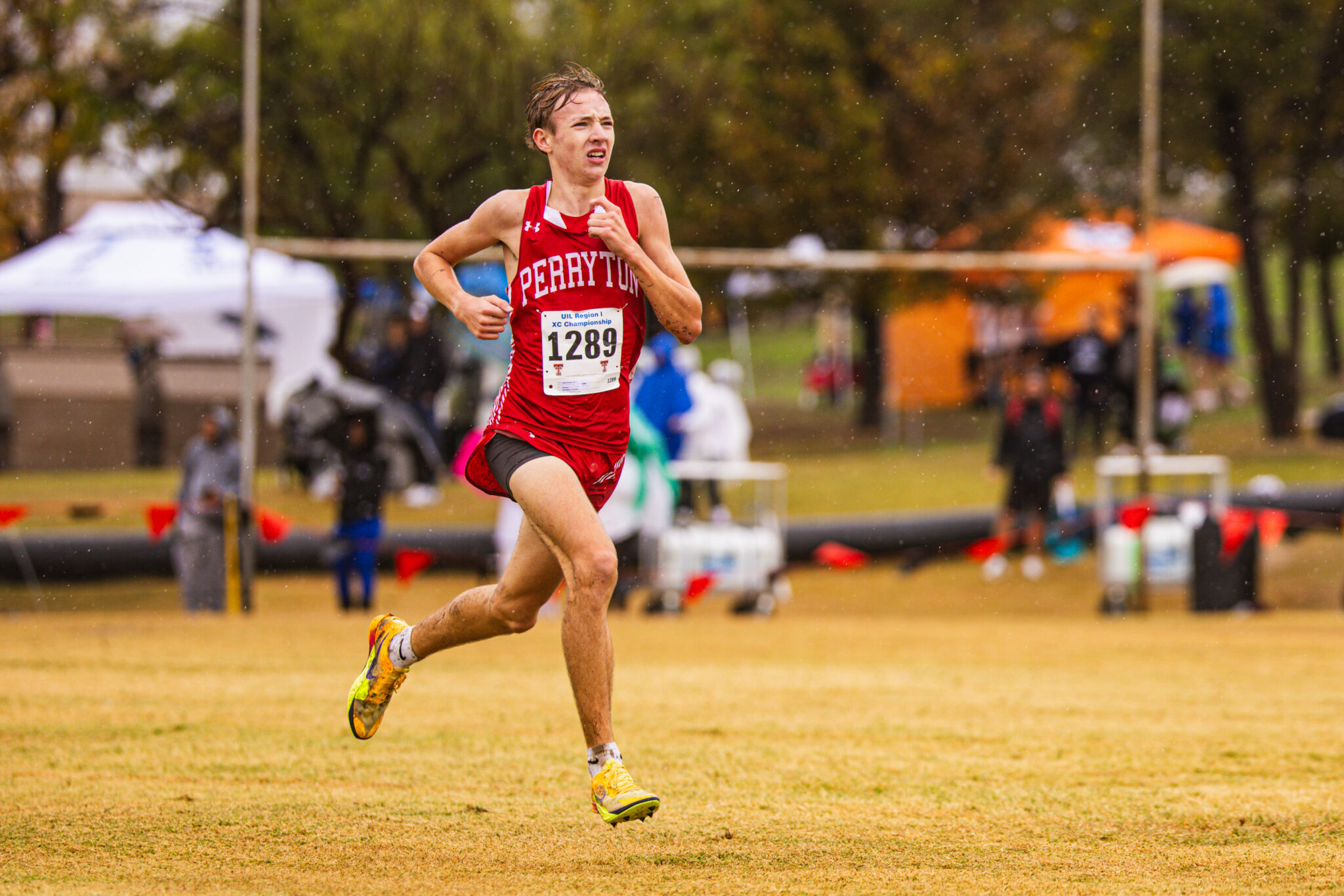 Male runner in a red racing kit with bib 1289 sprinting on a wet, grassy field; raindrops visible in the air with blurred spectators in the background.