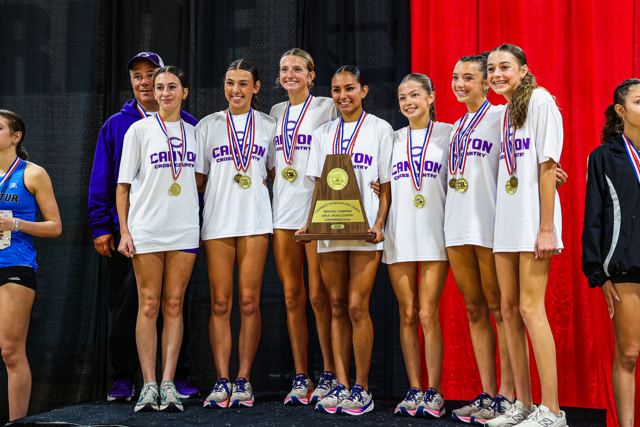Girls' cross-country team on stage wearing white shirts and medals, posing with a wooden trophy after a win.