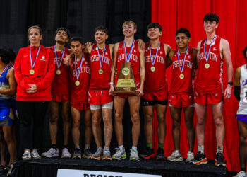 DEP23354 | Press Pass Sports Team of young runners in red uniforms with medals around their necks pose on stage, holding a wooden championship trophy in front of a red curtain backdrop.