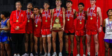 Team of young runners in red uniforms with medals around their necks pose on stage, holding a wooden championship trophy in front of a red curtain backdrop.