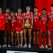 DEP23354 | Press Pass Sports Team of young runners in red uniforms with medals around their necks pose on stage, holding a wooden championship trophy in front of a red curtain backdrop.