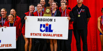Group of young athletes on a stage in red and black jackets, smiling with medals, holding a trophy and a Region 1/State Qualifier banner against red curtains and a black backdrop.