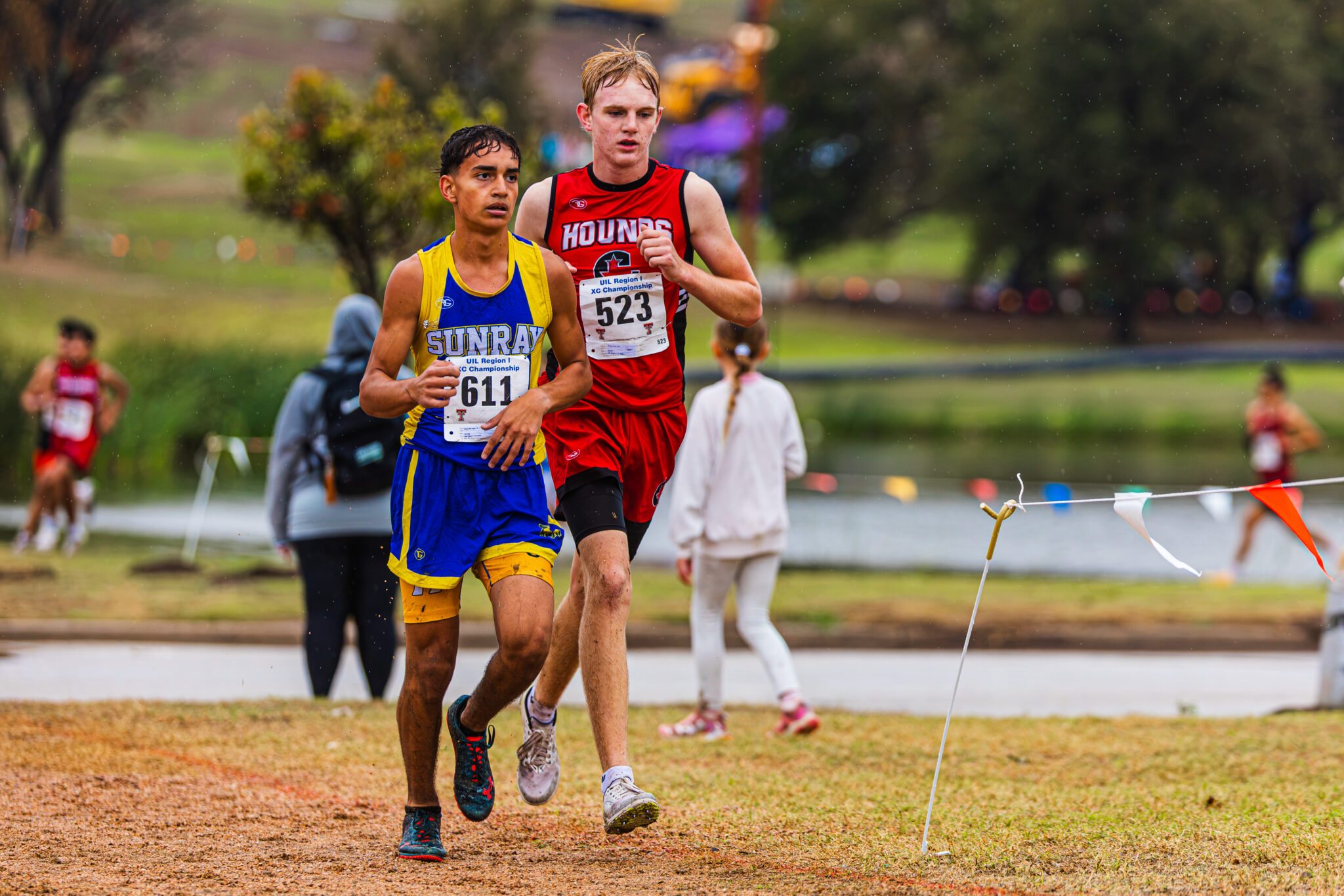 DE3_7884 | Press Pass Sports Two young male runners in a cross-country race, wearing blue-yellow and red uniforms with bibs 611 and 523, sprint on a dirt path in the rain.