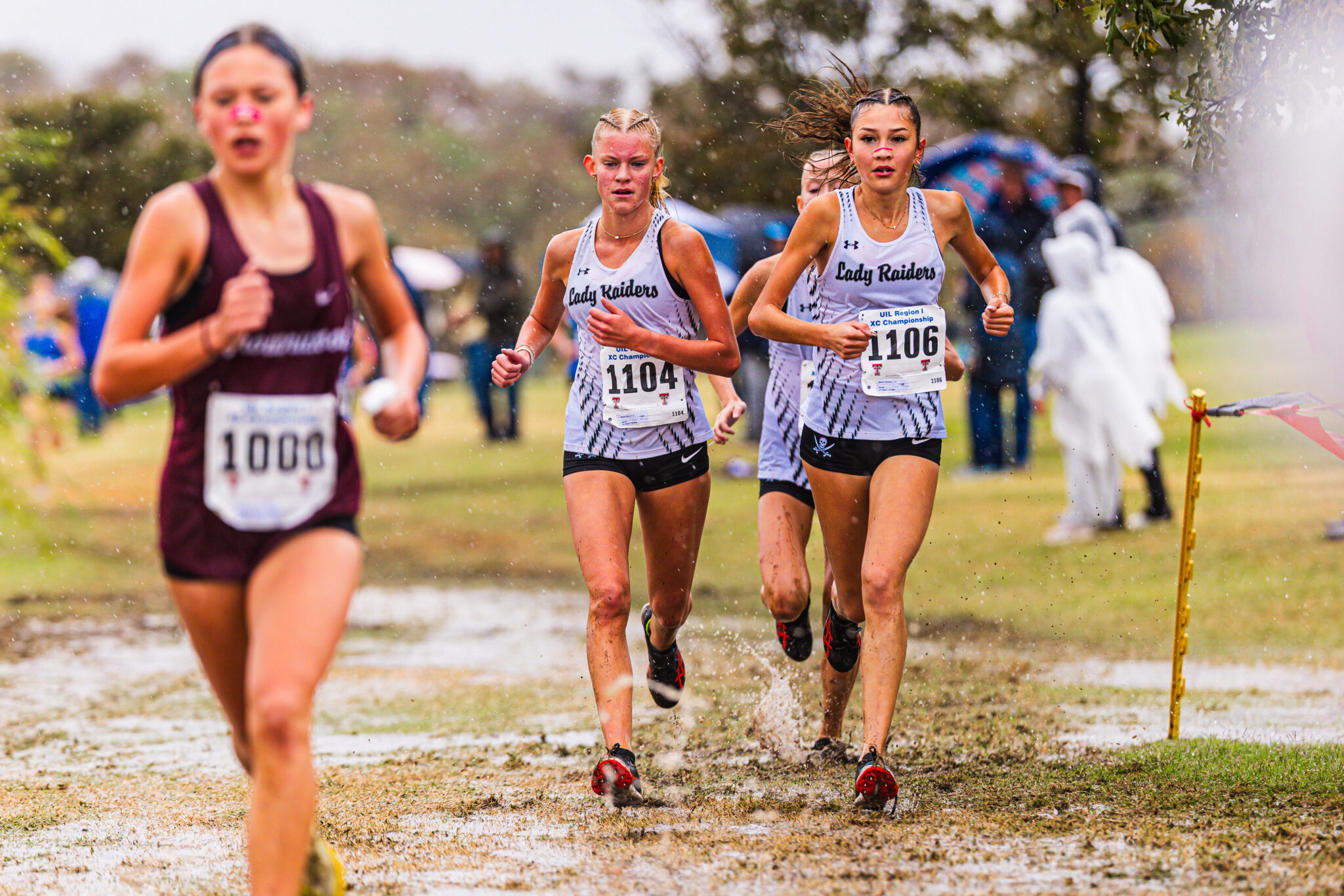 Young female runners sprint through a muddy cross-country course; two in white'Lady Raiders' jerseys lead, bib numbers 1104 and 1106 visible.