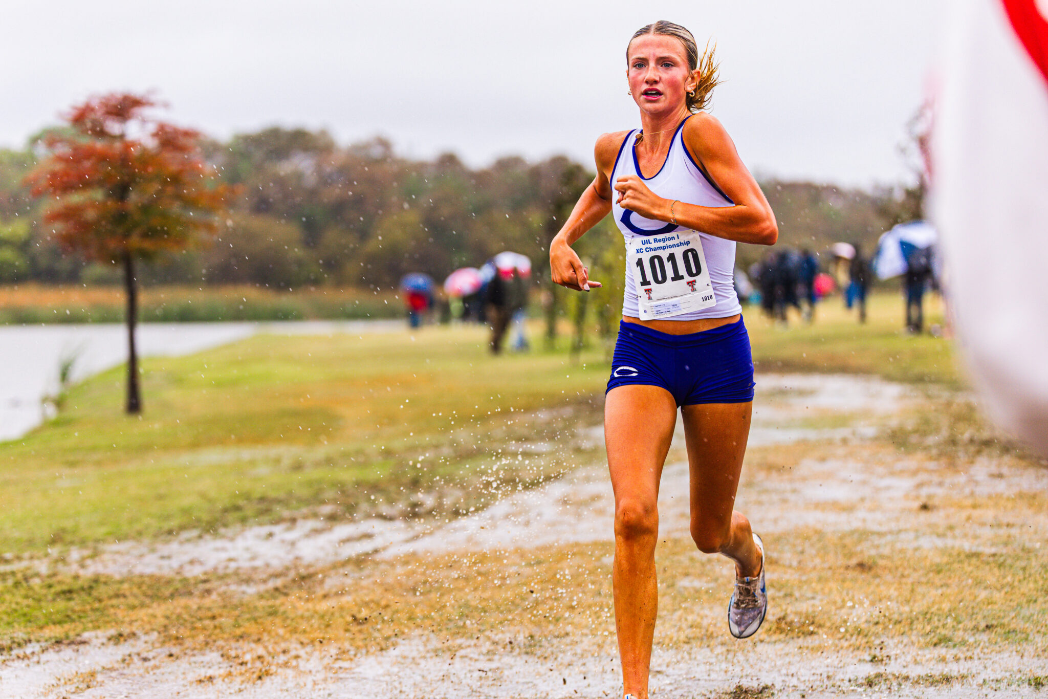 Female runner in white and blue, bib 1010, sprinting on a wet, muddy course during a race.