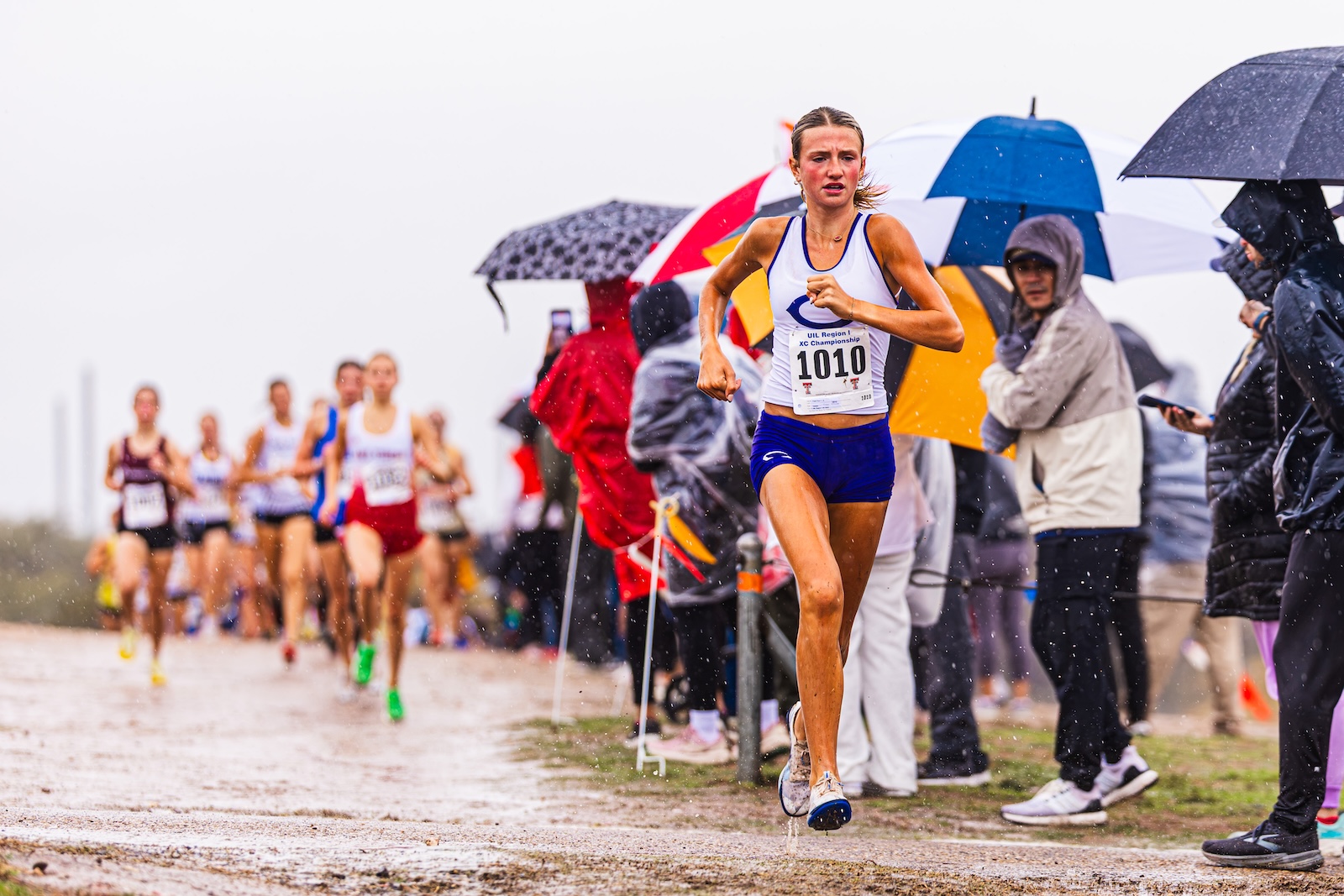 Female runner with bib 1010 leading a rain-soaked race along a muddy path, spectators under umbrellas on the sidelines.