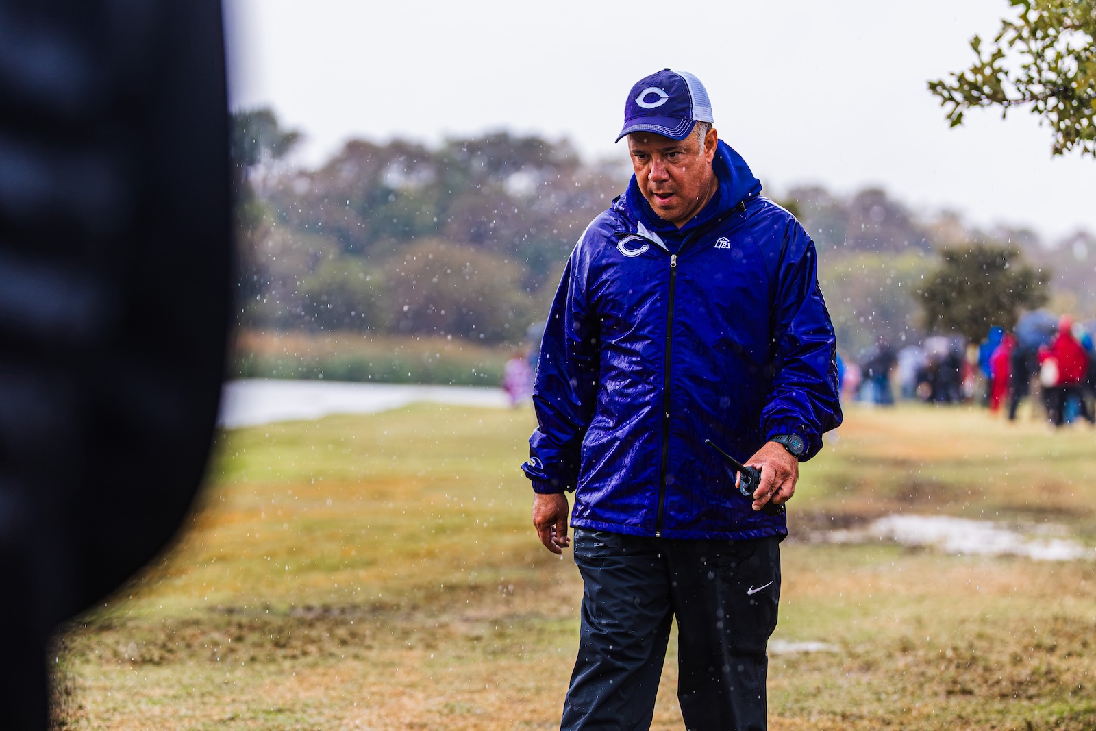 Man in a blue rain jacket and cap standing in a rainy park, holding a walkie-talkie amid a crowd in the background to the right.