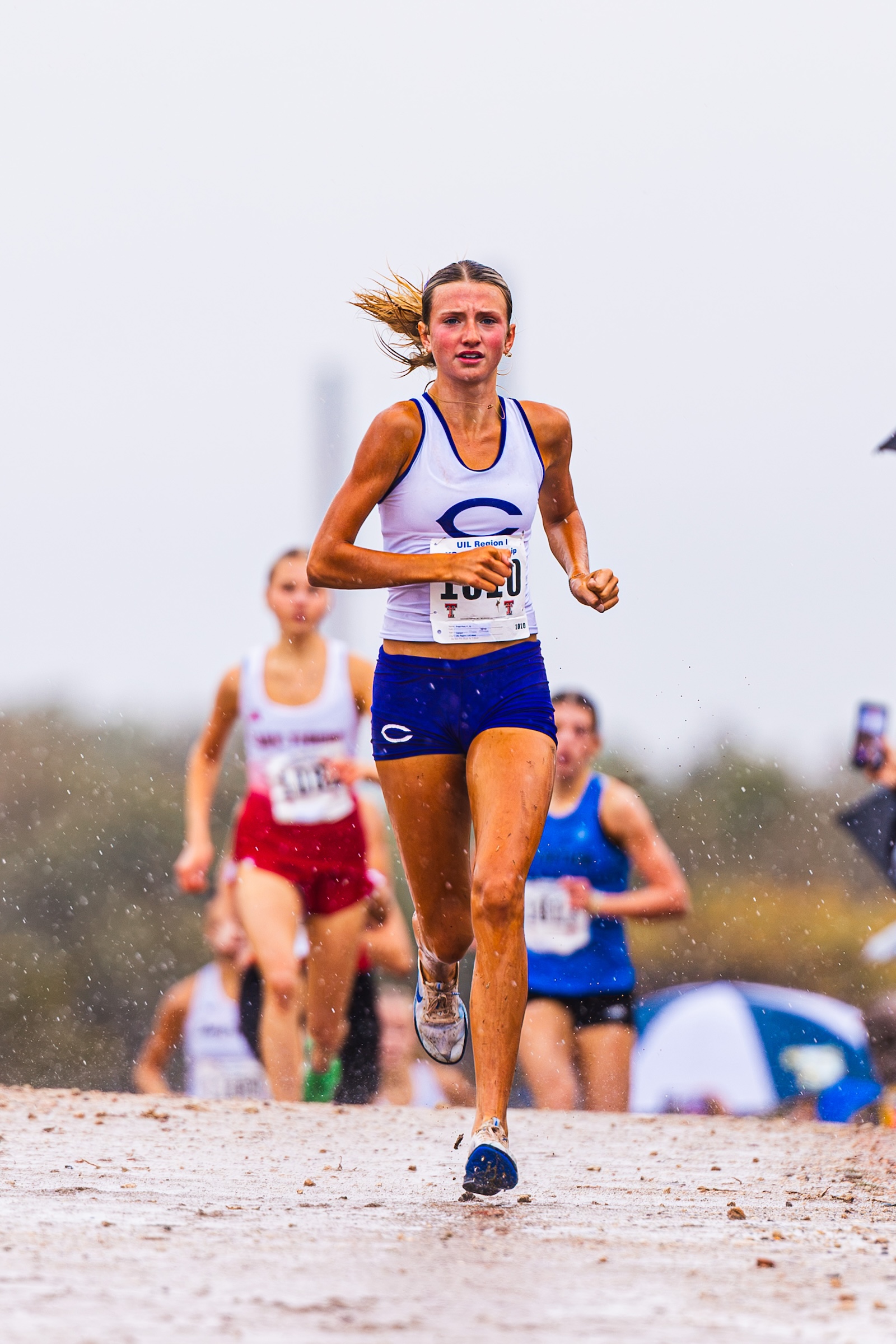 Female runner in a white and blue kit leads the race on a sandy track, with competitors behind her.