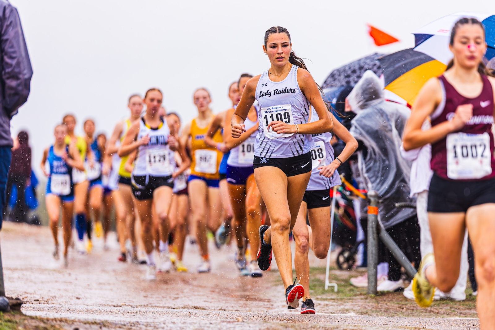 Female runner in a gray 'Lady Raiders' jersey leads a rain-soaked race, with a crowded pack behind on a muddy path.