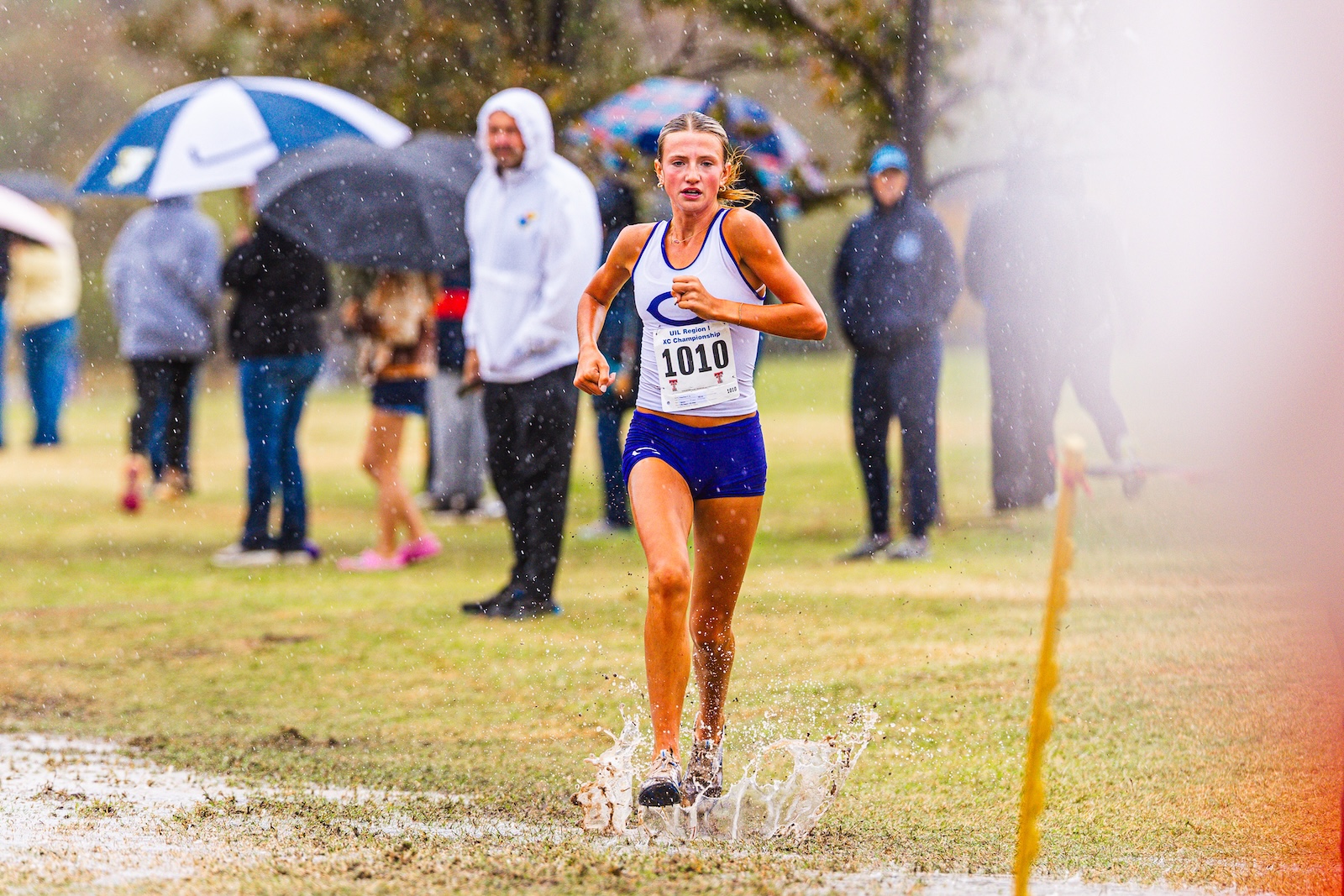Female runner wearing blue shorts and white tank top sprinting through a muddy puddle during a rainy cross-country race, spectators with umbrellas in the background.