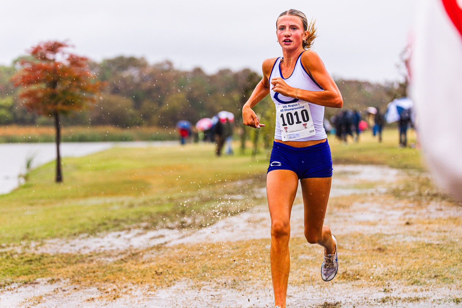 Female runner in a white tank top and blue shorts with bib 1010, sprinting on a muddy path beside a lake with spectators in the background.