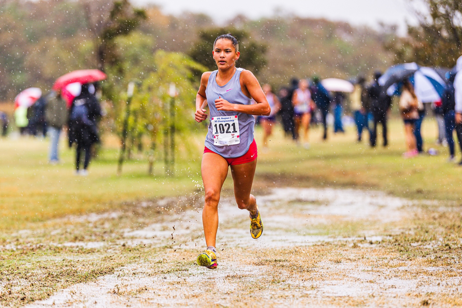 Female runner in gray tank top and red shorts sprinting on a muddy cross-country path, bib 1021 visible, spectators with umbrellas in the background.