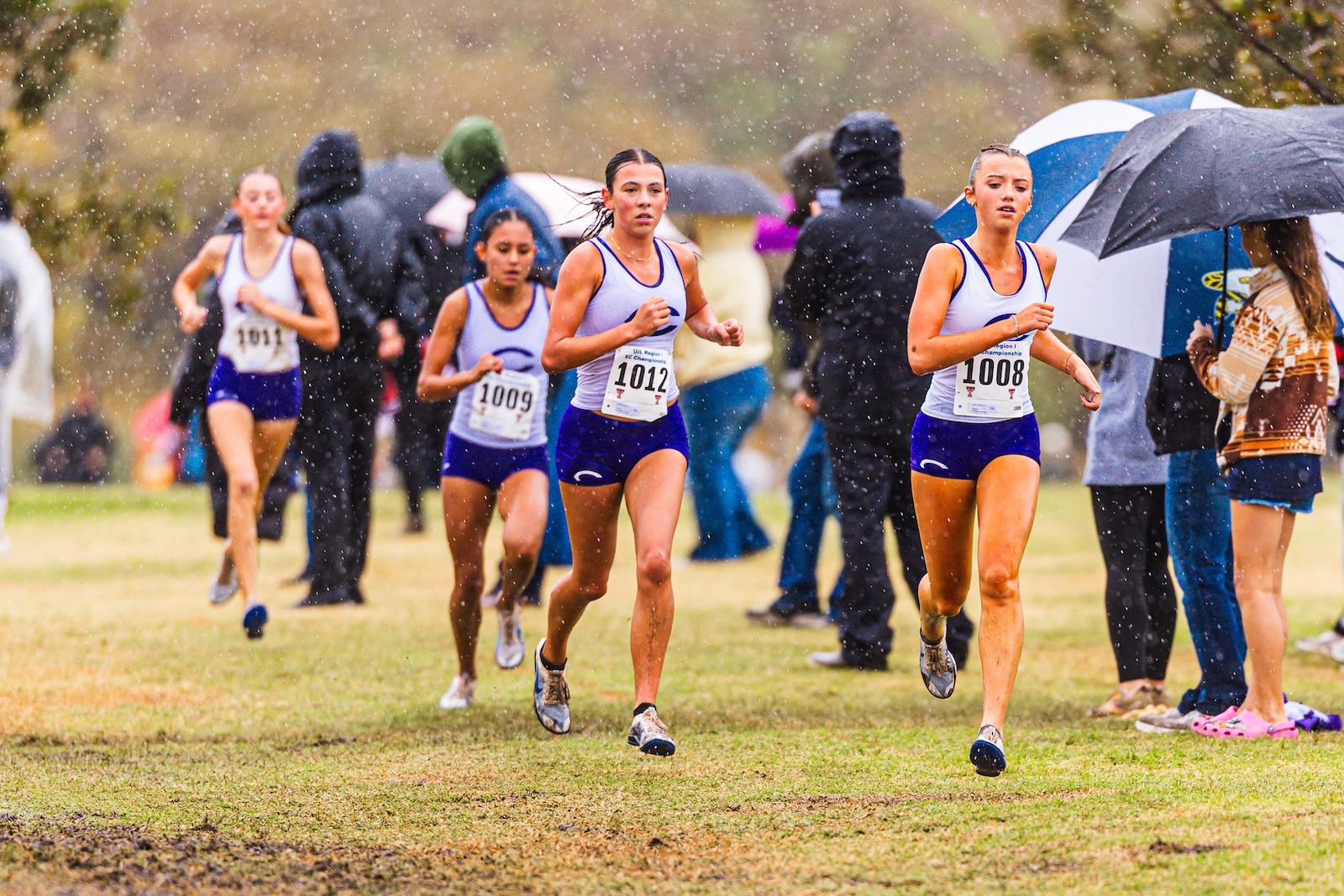 Female runners in purple uniforms sprint on a rainy cross-country course while spectators with umbrellas watch nearby.