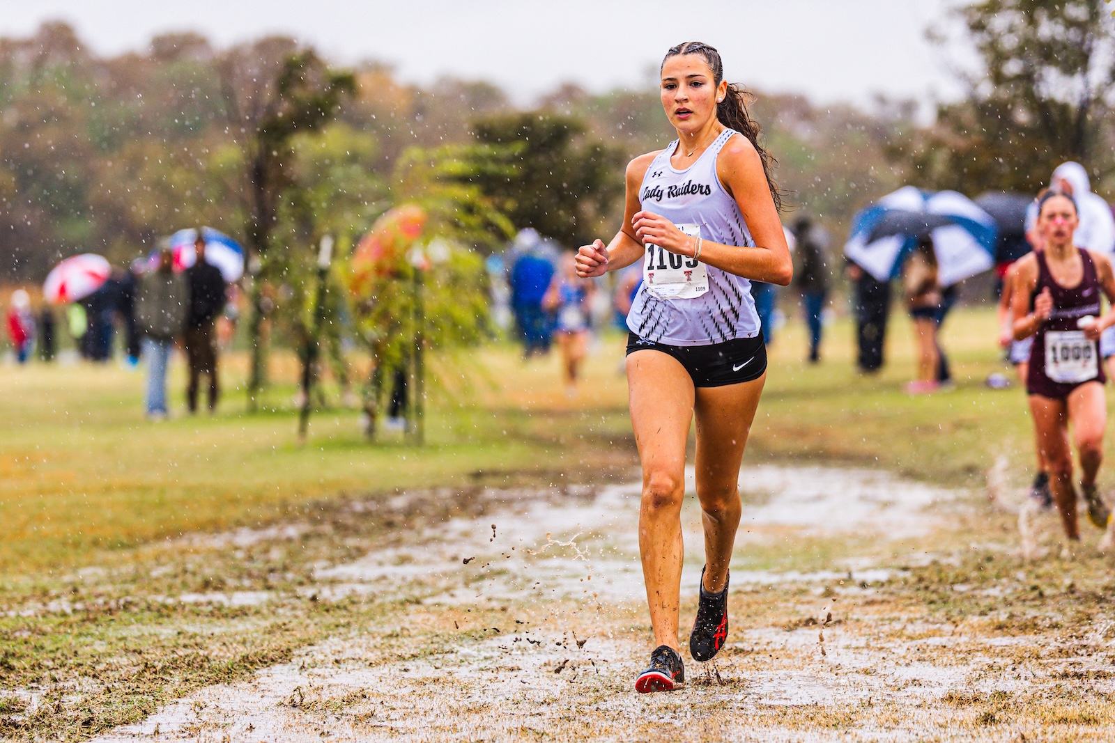 Female runner in a light blue racing singlet and black shorts moves through a muddy, rain-soaked cross-country course, bib number visible.