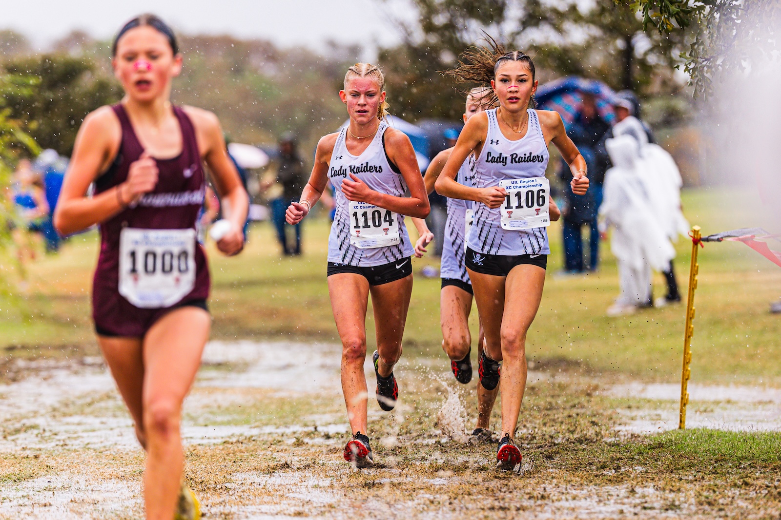 Girls' cross-country race on a muddy field; three runners in white jerseys labeled 'Lady Raiders' with bib numbers 1104 and 1106 leading, kicking up water as they run.