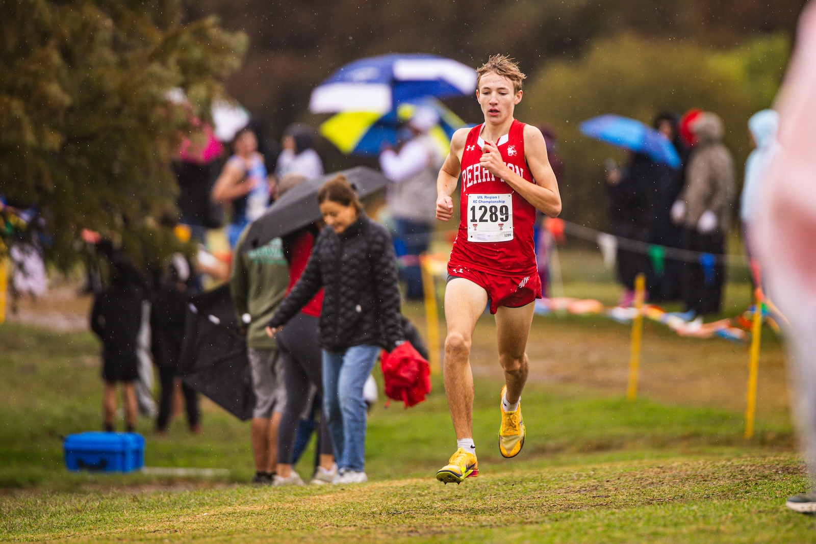 Male cross-country runner in a red uniform sprinting on a muddy track, bib 1289, with spectators under umbrellas in the rain behind him.