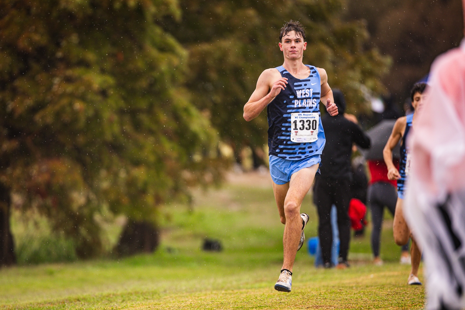 Male runner in a blue West Plains singlet with bib 1330, sprinting on a grassy cross-country course in light rain.