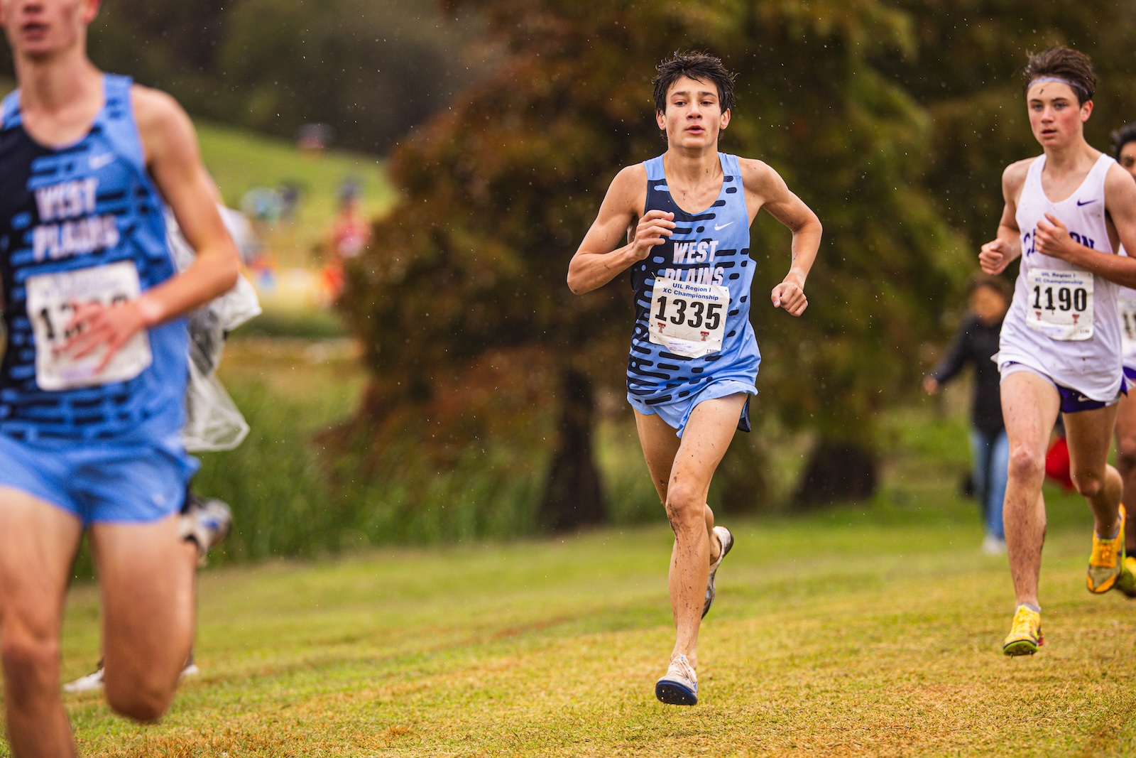 Young male runner in a blue West Plains singlet with bib 1335 leading a cross-country race in wet grass.