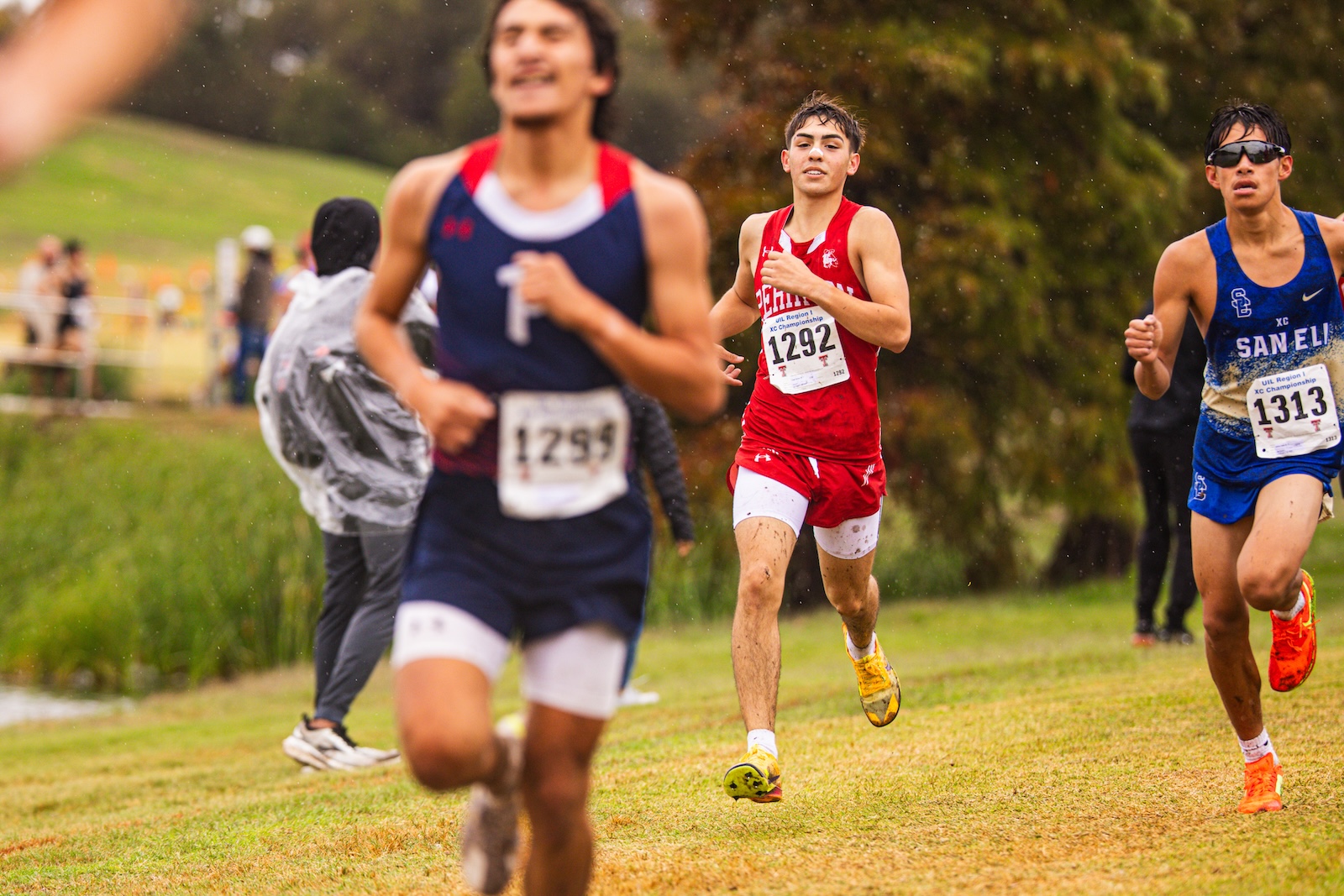 Young runners sprint on a grassy cross-country course, wearing red and blue uniforms and numbered bibs, in light rain.