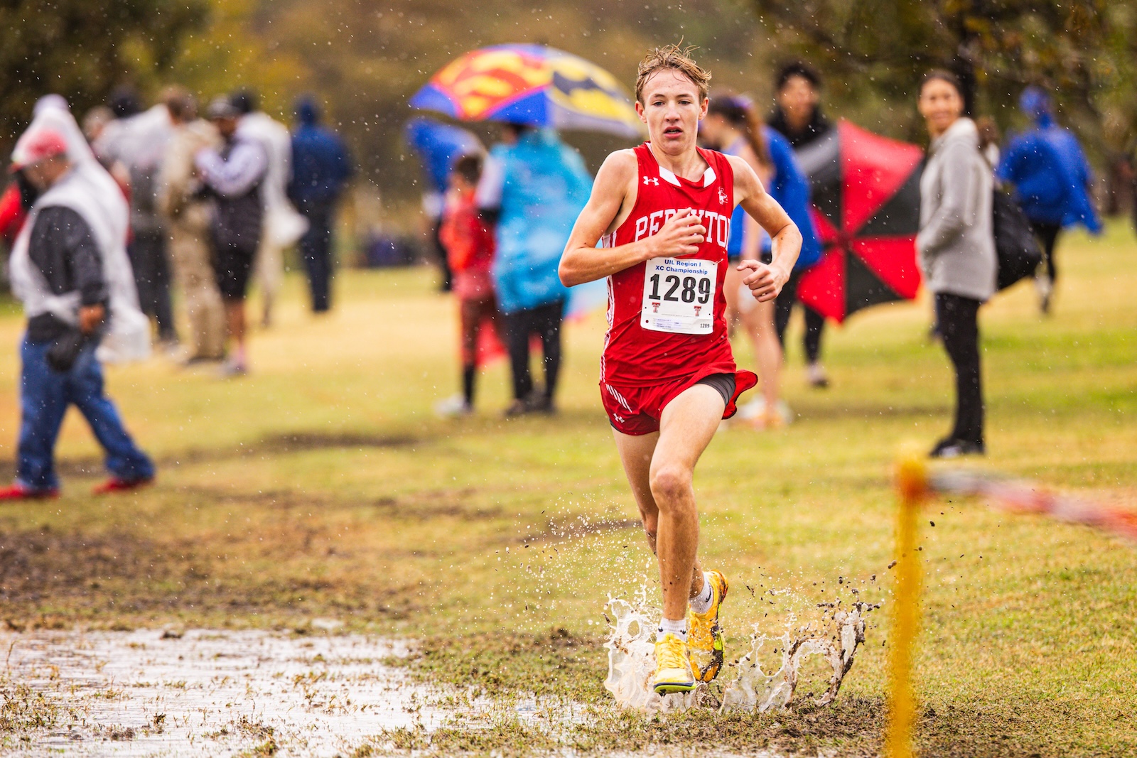 Male cross-country runner in a red uniform splashes through a muddy puddle as spectators watch in the rain with umbrellas behind him.