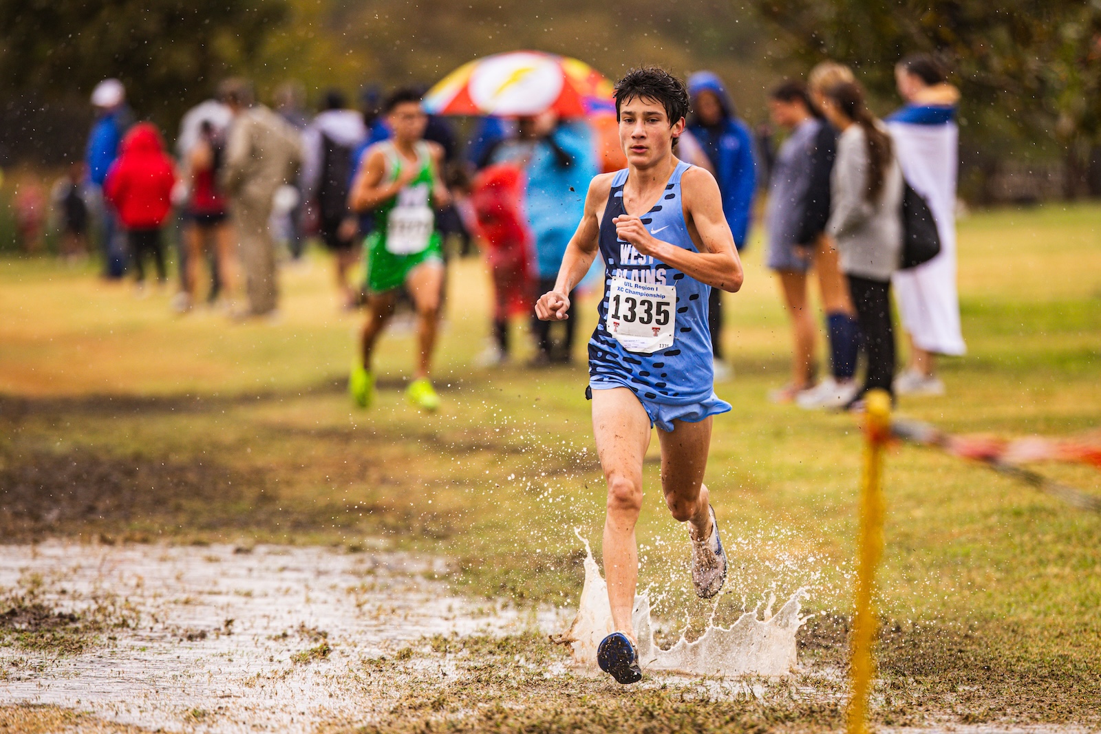 Runner in blue singlet and shorts splashes through a muddy puddle during a cross-country race, bib 1335, with spectators blurred in background.