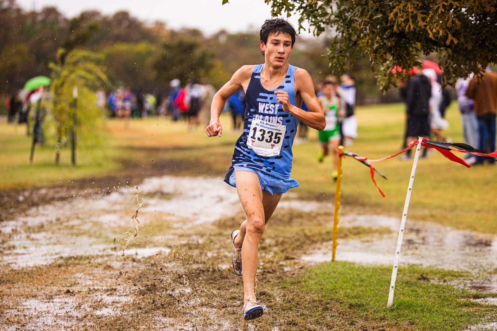 Young male runner in a blue singlet and shorts sprinting through a muddy cross-country course, bib 1335, spectators in the background.