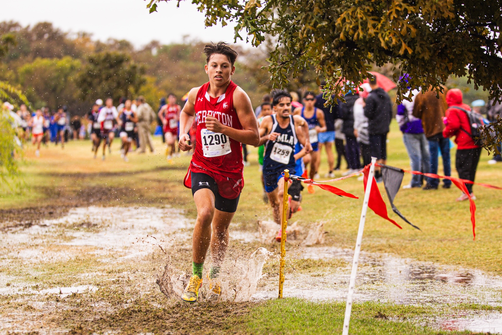 Front-running cross-country athlete in a red uniform splashes through a muddy puddle as spectators watch in the background.