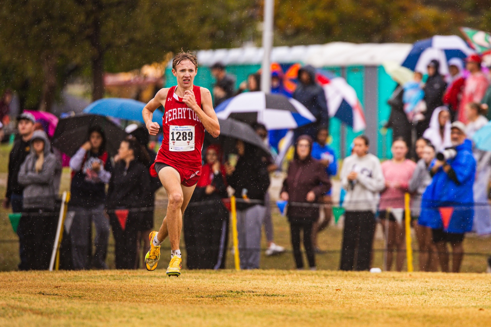 Runner in a red singlet with bib 1289 sprinting on a rainy grassy field as spectators with umbrellas watch in the background.