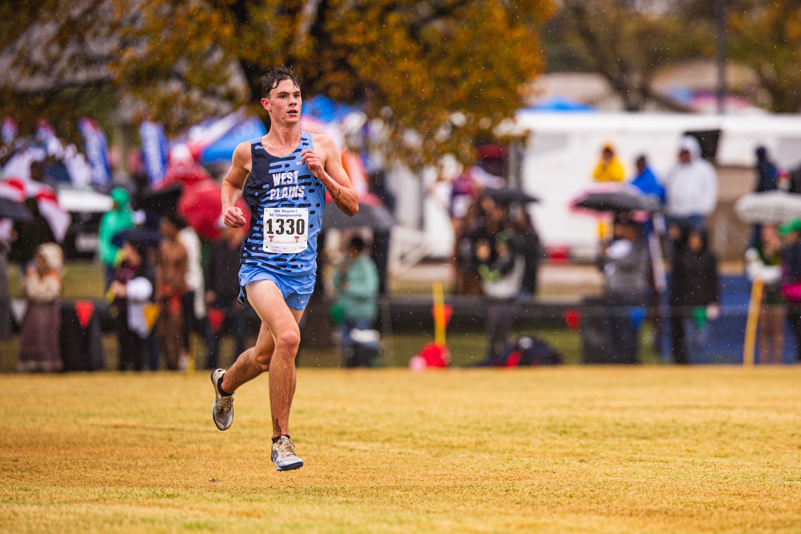 Runner in blue athletic kit with bib 1330 racing on a grassy field, spectators in the background on a rainy day.