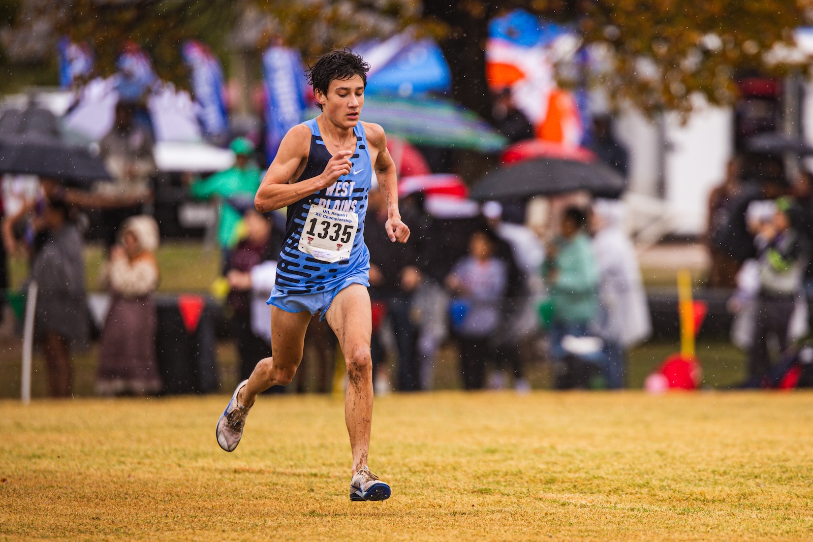 Male runner in a blue uniform with bib 1335 sprinting on a muddy field in the rain, with spectators behind him.