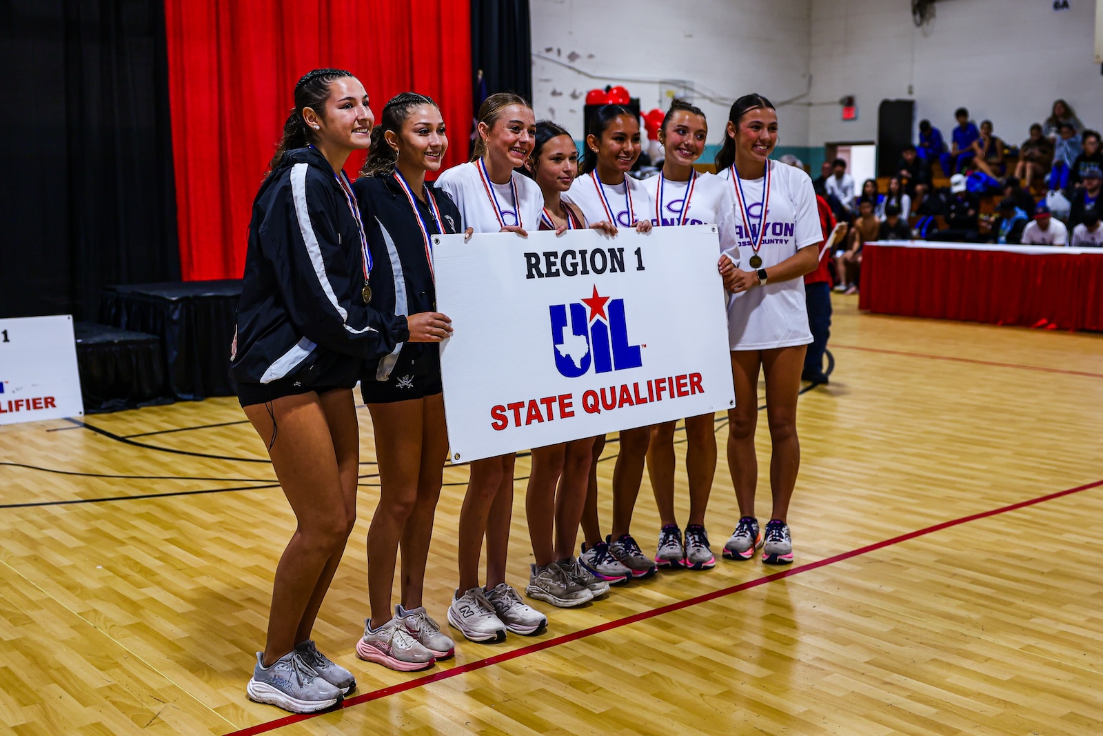 Young girls in athletic gear posing with a UIL State Qualifier banner and medals after a competition in a gymnasium gym.