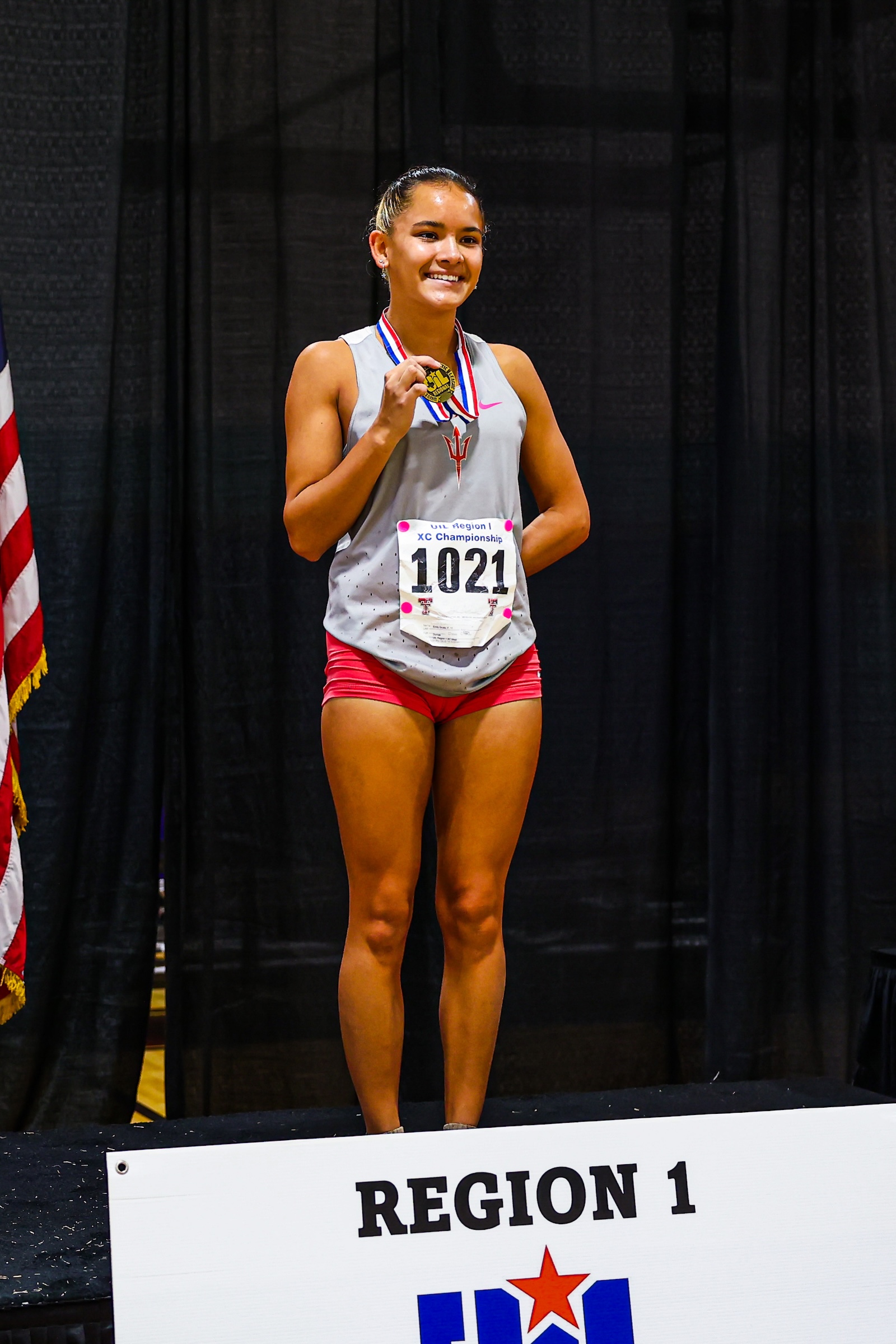 Young female athlete on a podium wearing a medal and gray top, red shorts, smiling at awards ceremony beside a 'Region 1' banner.