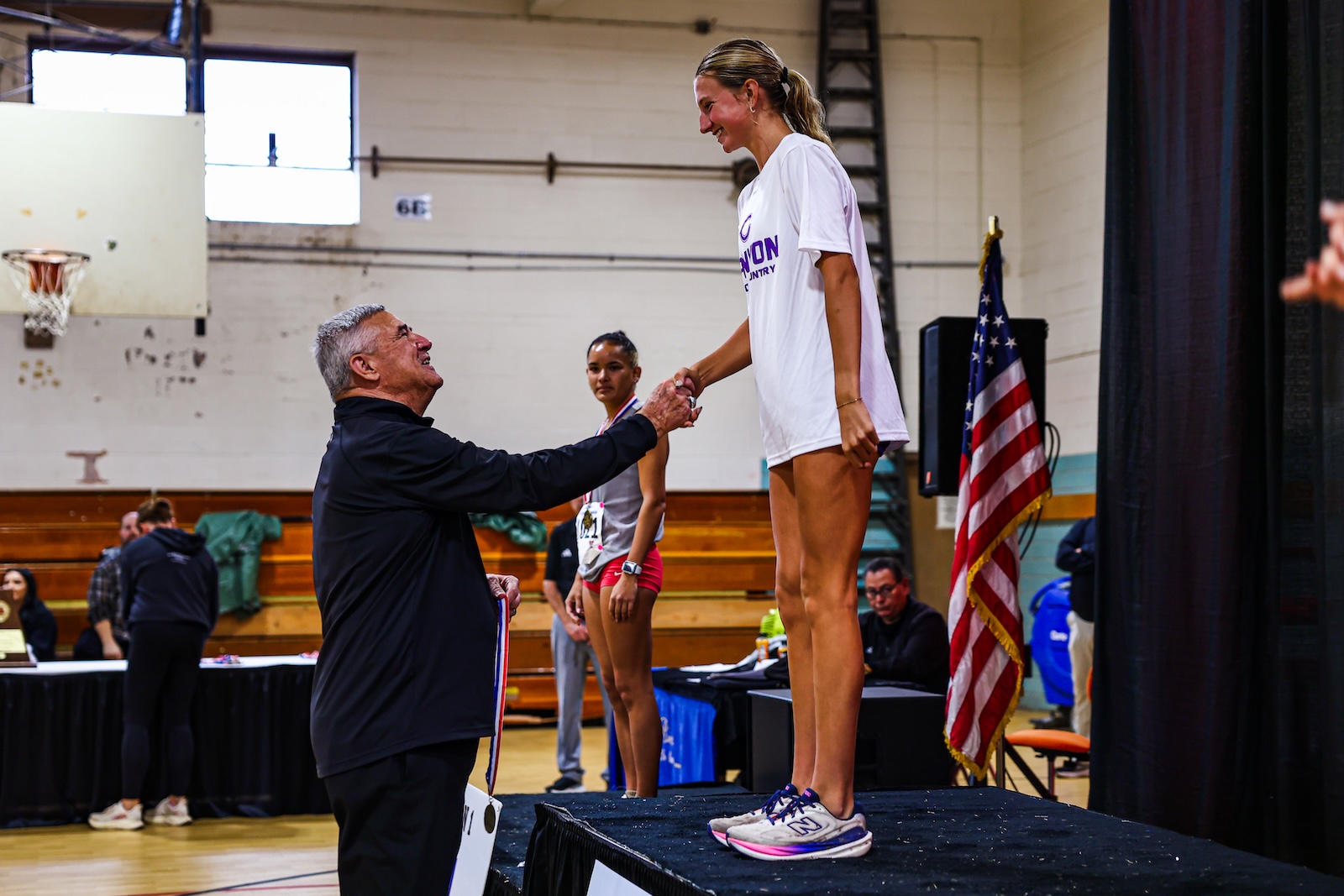 Coach hands a medal to a tall smiling athlete on a stage in a gym, with an American flag in the background.