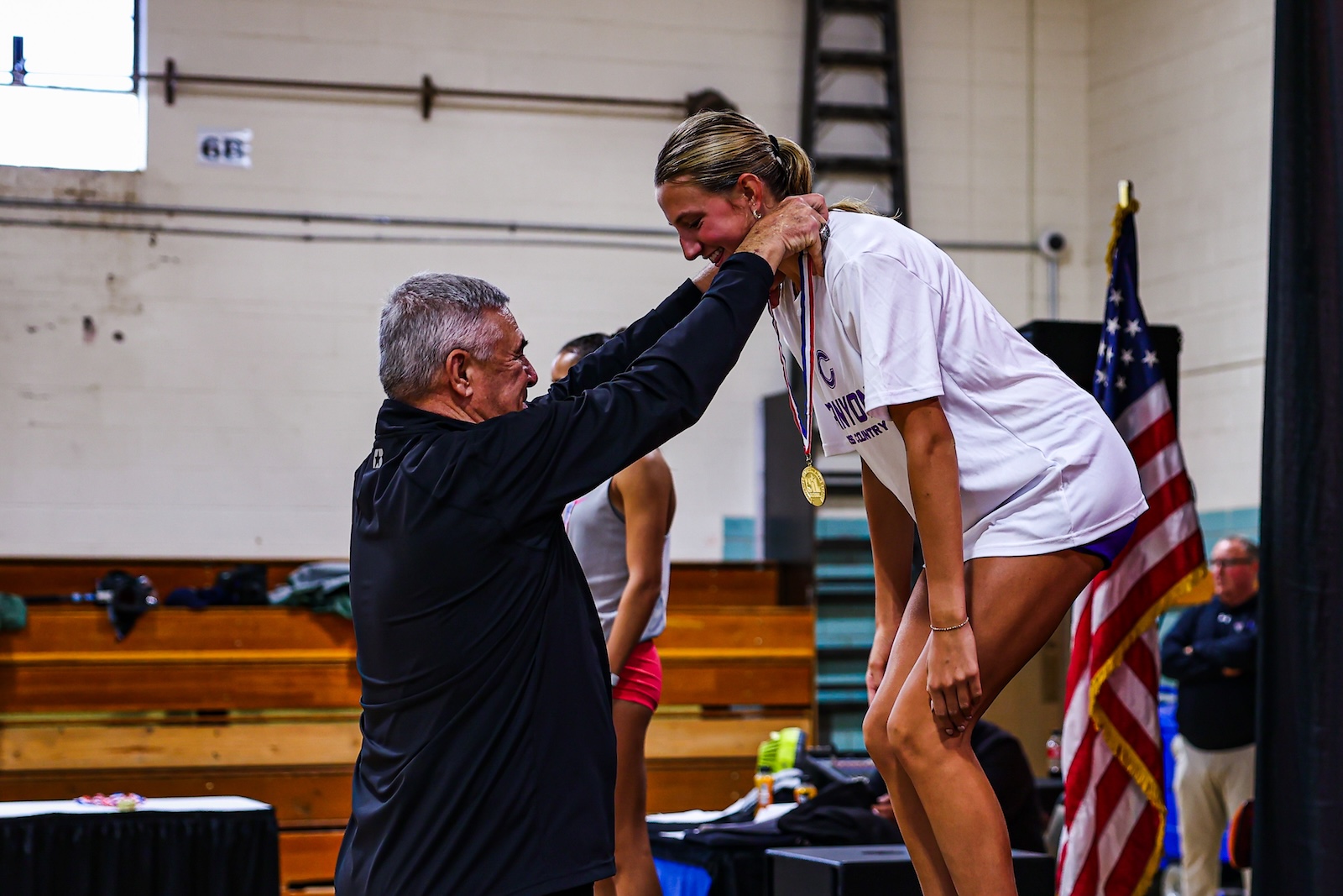 Coach medals a female athlete as she bows on a podium, gold medal hanging from her neck in a gymnasium with an American flag nearby.