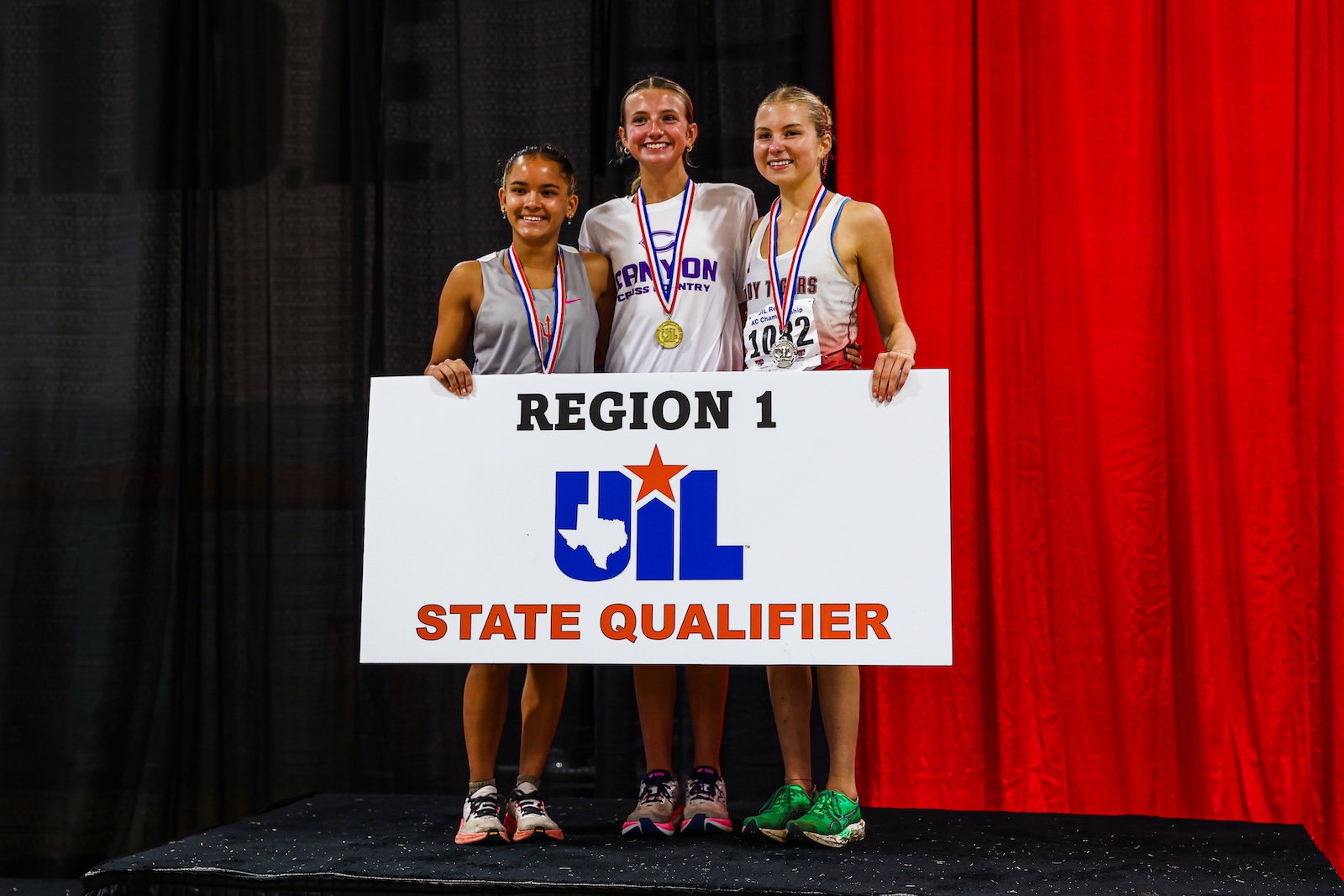 Three smiling runners stand on a podium holding a large sign that reads REGION 1 UIL STATE QUALIFIER, medals around their necks, red curtain backdrop.