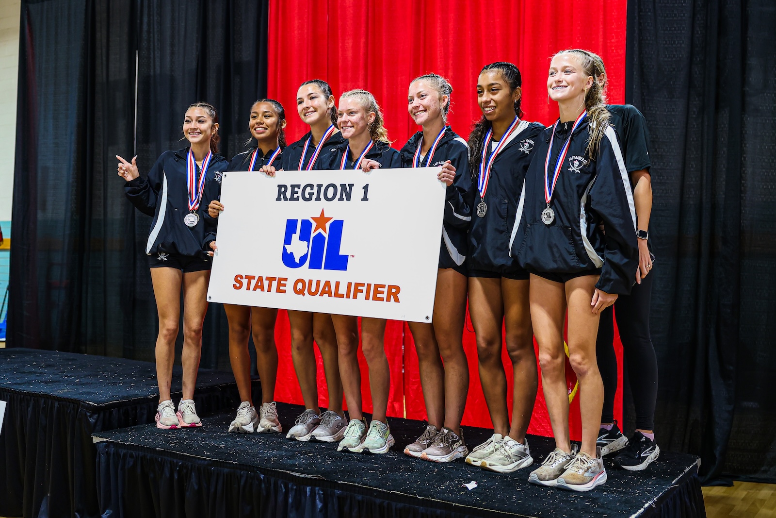 Girls' athletic team on stage posing with a UIL State Qualifier sign and medals.