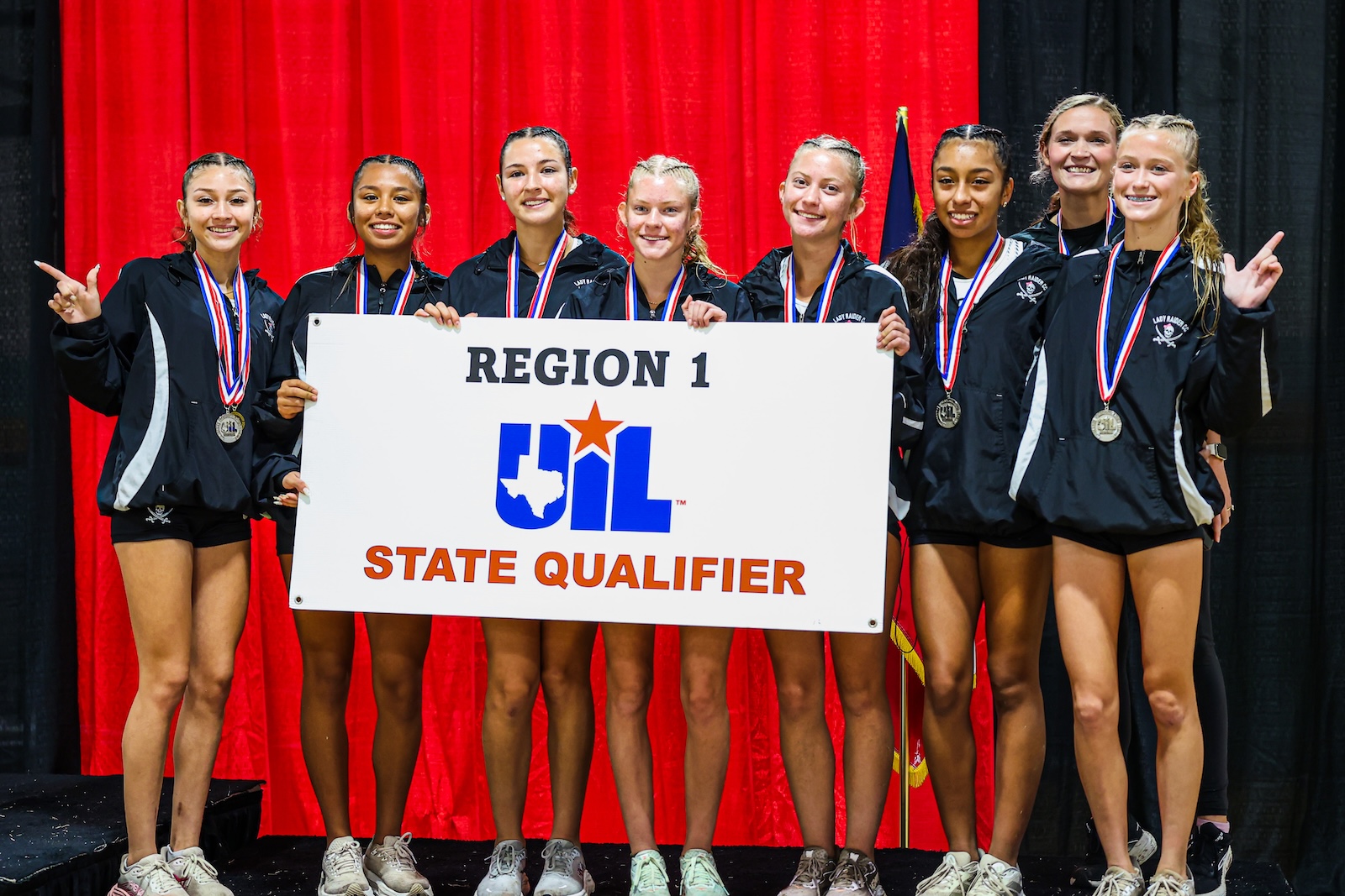 Girls' high school team on stage holding a UIL State Qualifier banner with medals, smiling after a competition.