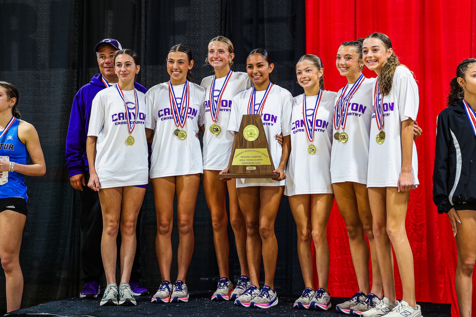 Girls' cross-country team in white shirts with medals, posing on stage with a large trophy center-stage.