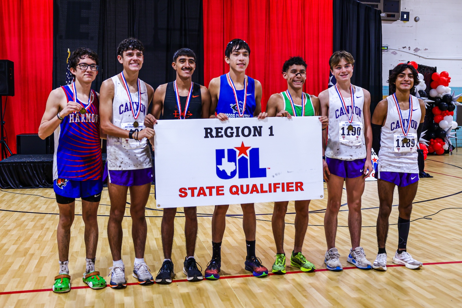 Seven boys in athletic uniforms with medals, holding a UIL Region 1 State Qualifier banner in a gym with red curtains behind them.