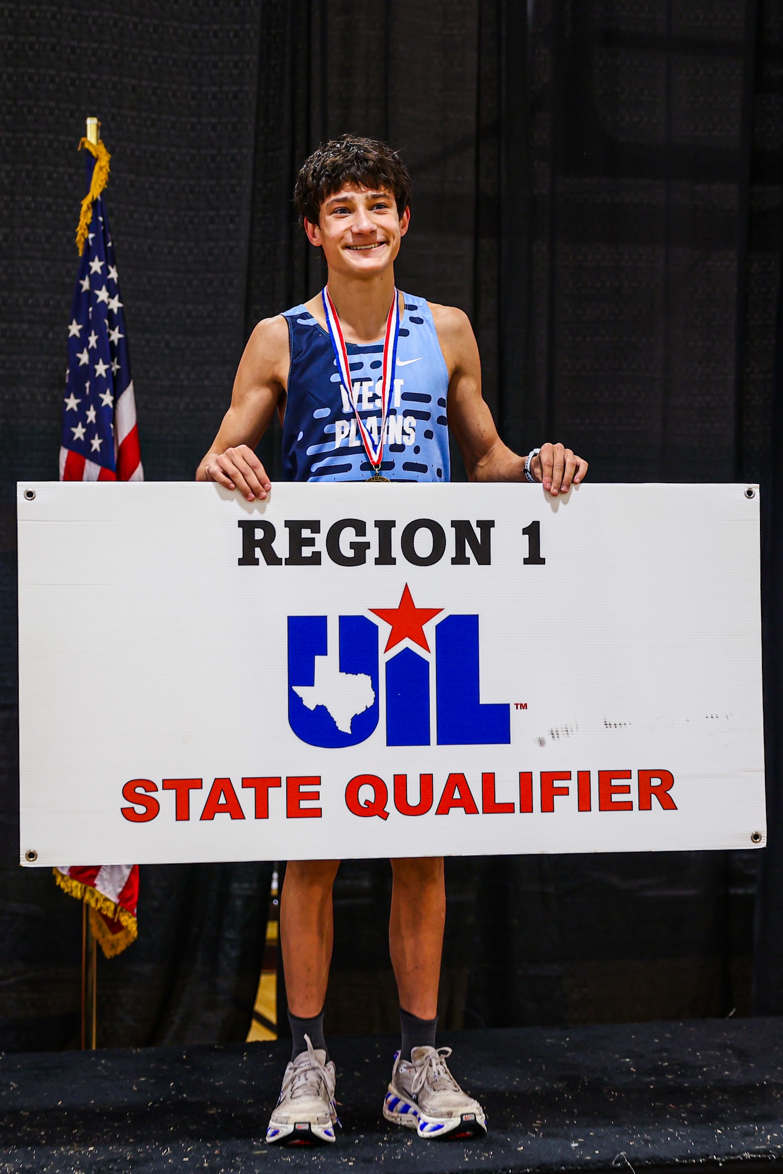 Smiling young runner on stage holding a Region 1 State Qualifier sign with a medal, American flag beside him.