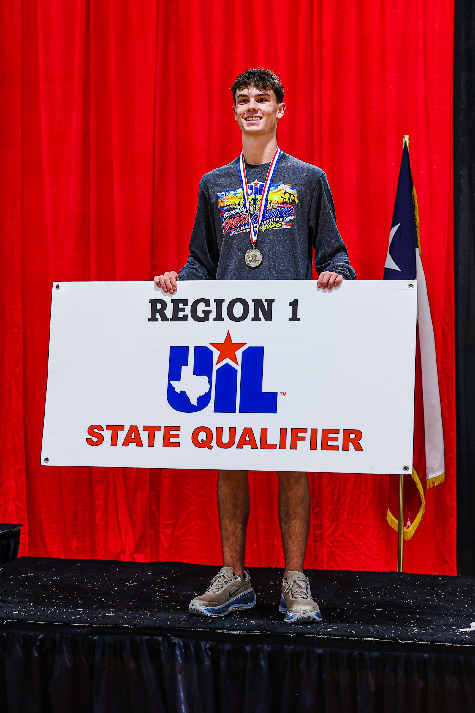 Young man on a stage wearing a medal, holding a large sign that says REGION 1 UIL STATE QUALIFIER, red curtain background.