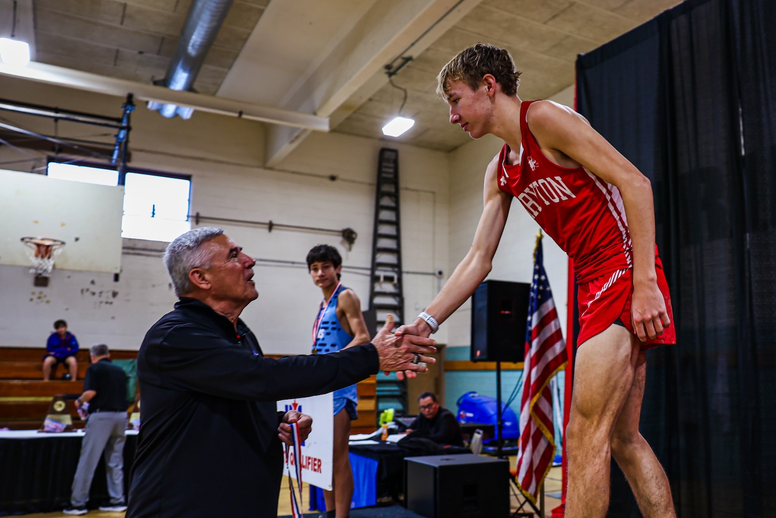 Wrestler in a red uniform shakes hands with an older man during an awards ceremony in a gym, medals on display and an American flag in the background.
