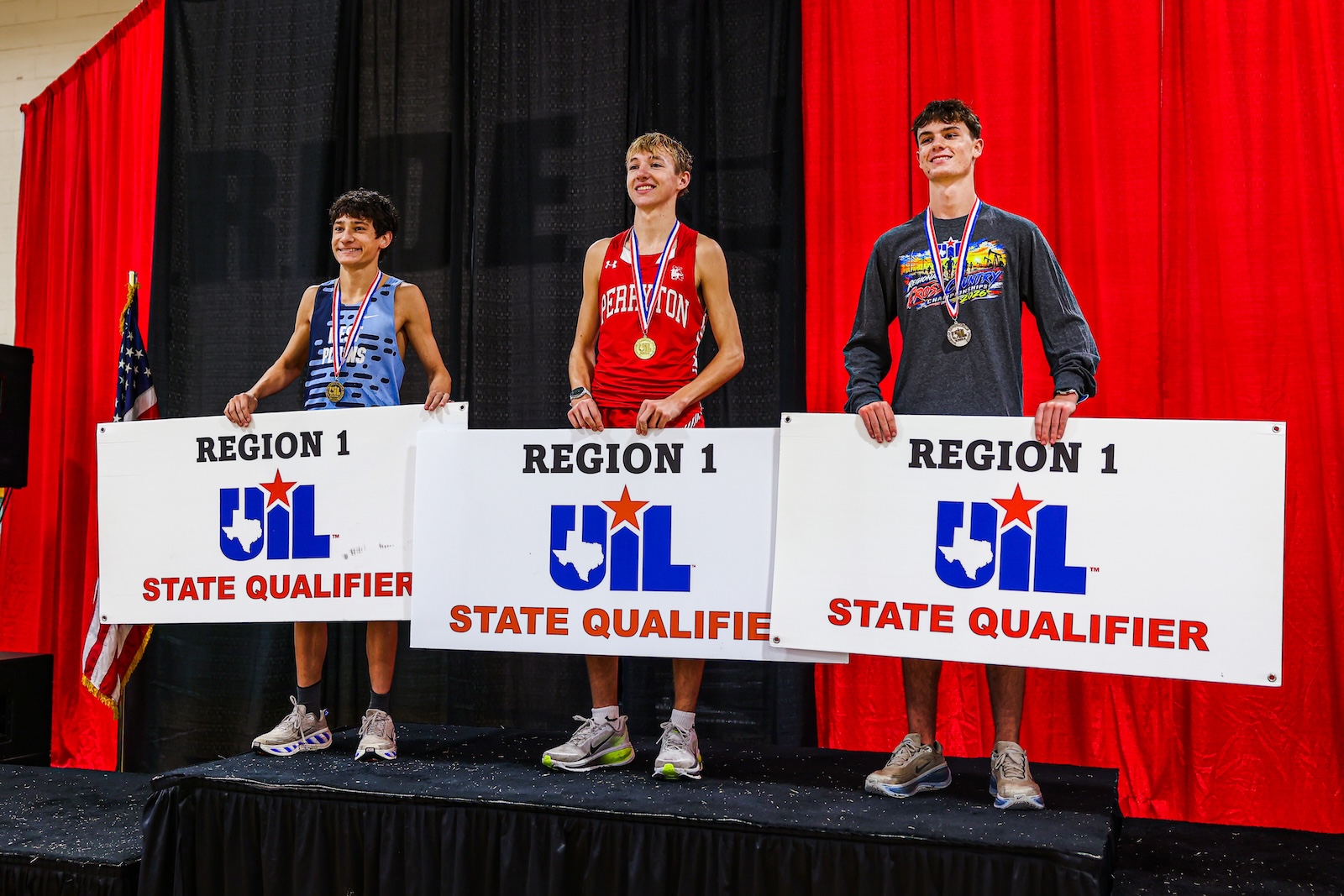 Three young male athletes stand on a podium holding Region 1 UIL State Qualifier signs, wearing medals on red, blue, and gray uniforms, with red curtains and an American flag nearby.