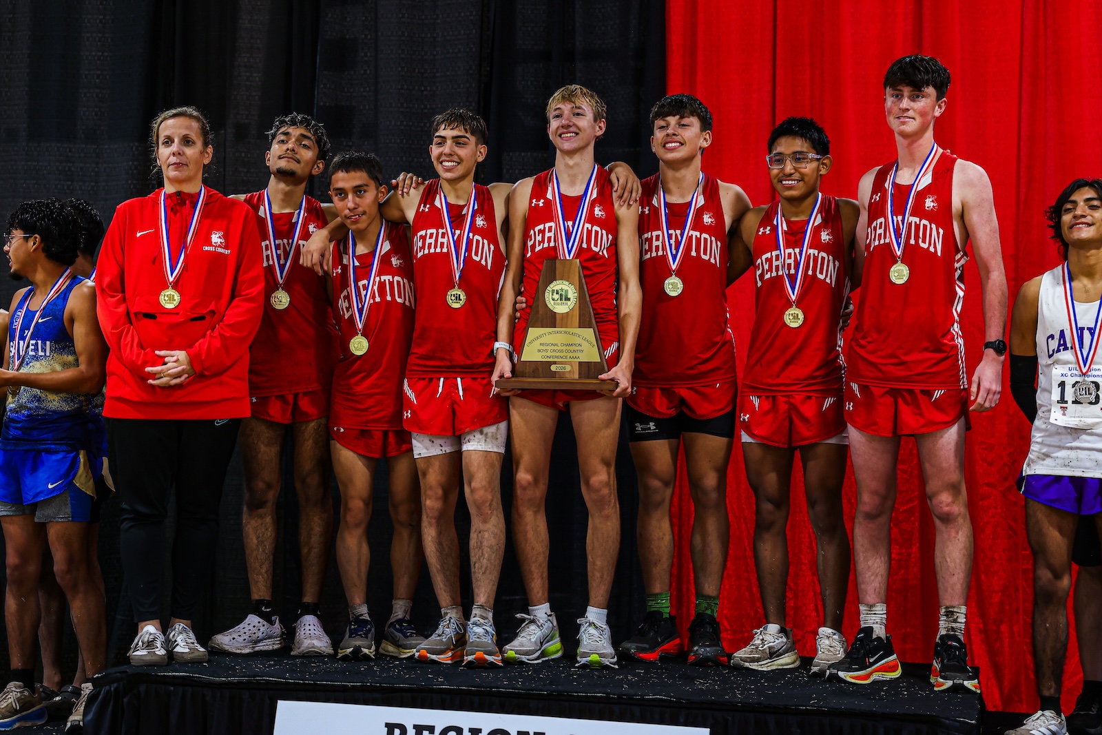Perriton boys' cross-country team on stage in red uniforms, wearing gold medals and posing with a large trophy against a red curtain backdrop.