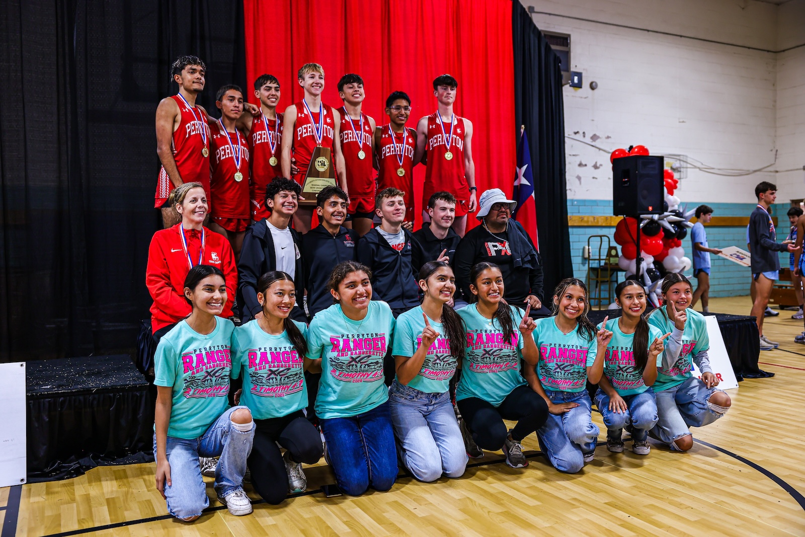 Group of young athletes posing on a stage after a sports event; front row in teal shirts, back rows in red uniforms with medals, trophy centered on stage.