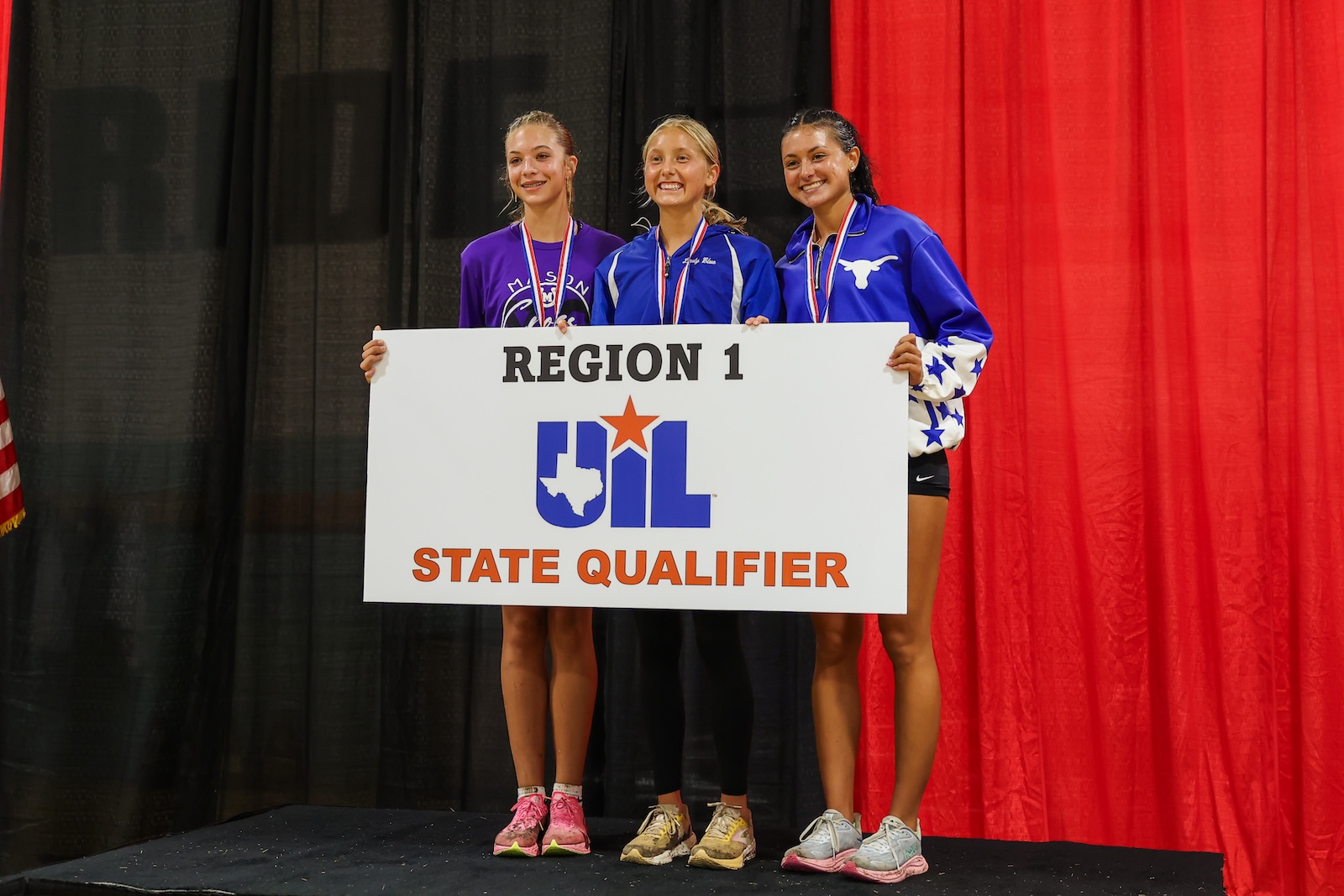 Three female athletes stand on stage holding a UIL State Qualifier sign, wearing medals against black and red curtains.