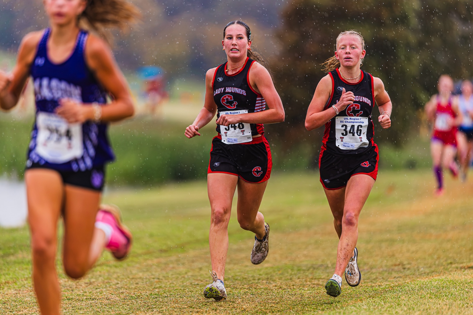 DE3_7152 | Press Pass Sports Two female cross-country runners in black and red uniforms ('Lady Hounds') lead a race on a damp grassy course, while others blur in the background in light rain.