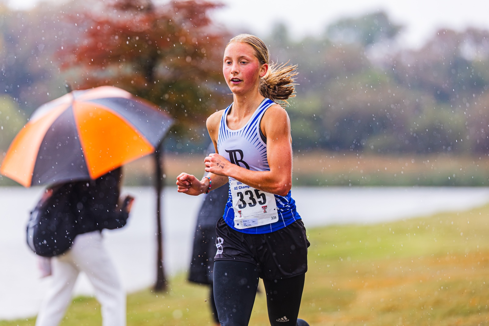 DE3_7296 | Press Pass Sports Female runner in a blue and white singlet with bib 335, sprinting in the rain along a park, with an orange umbrella in the background.